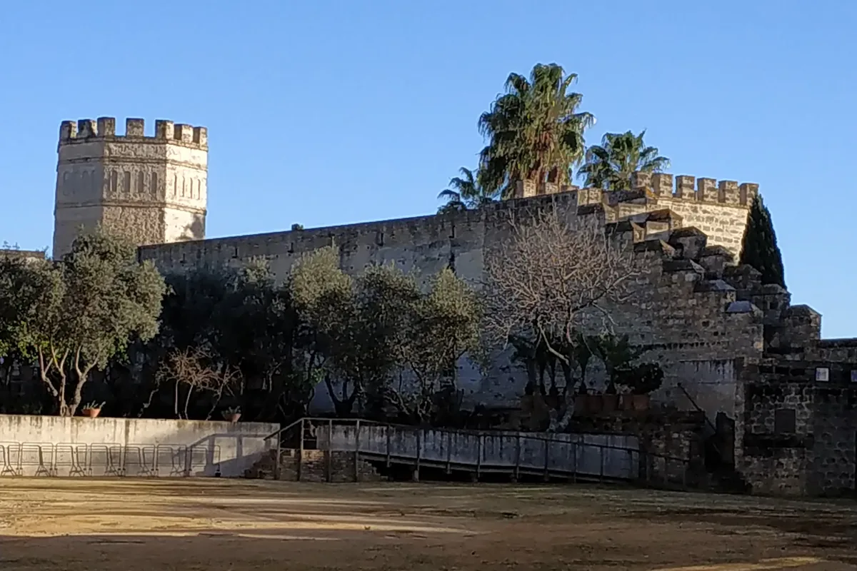 Ένα επάλξεις φέρων πέτρινο τείχος και ένας οκταγωνικός πύργος του Alcázar της Jerez που υψώνονται πάνω από ελιές και μια αμμώδη αυλή